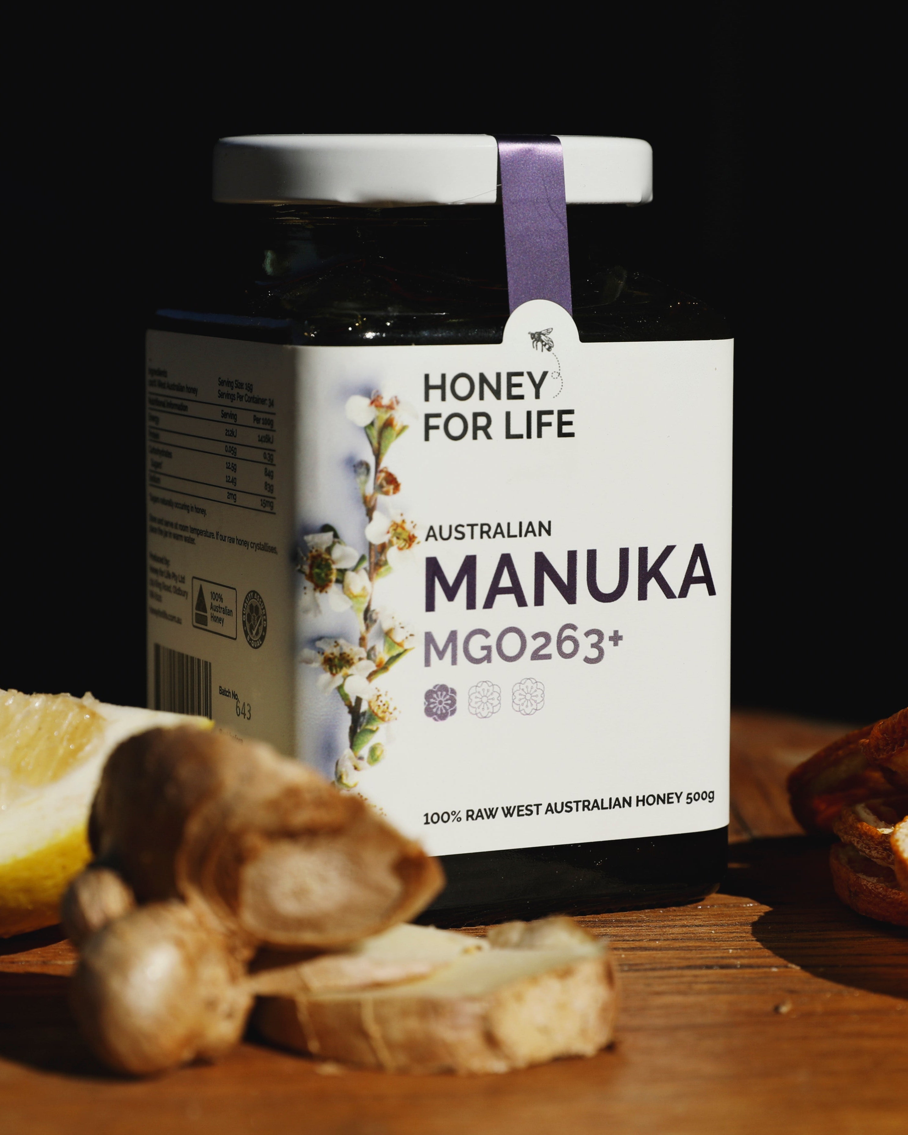 A jar of Australian Manuka honey labeled 'HONEY FOR LIFE' with MGO263+ indicated on the label. The jar is accompanied by slices of lemon and ginger on a wooden surface.