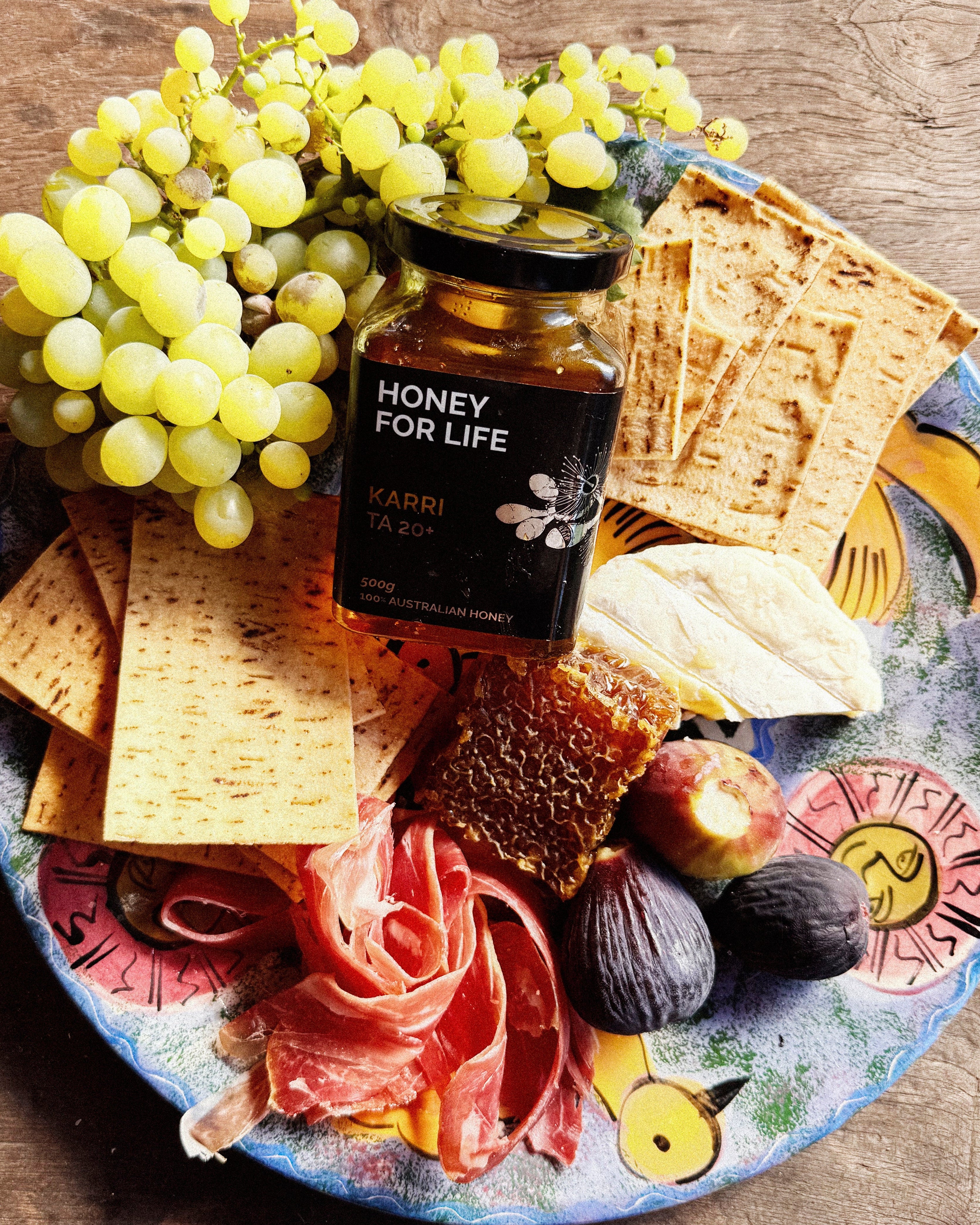 Honey for Life jar on a charcuterie board with grapes, figs, and crackers.