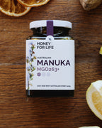 A jar of Australian Manuka honey labeled 'HONEY FOR LIFE' with MGO263+ indicated on the label. The jar is accompanied by slices of lemon and ginger on a wooden surface.