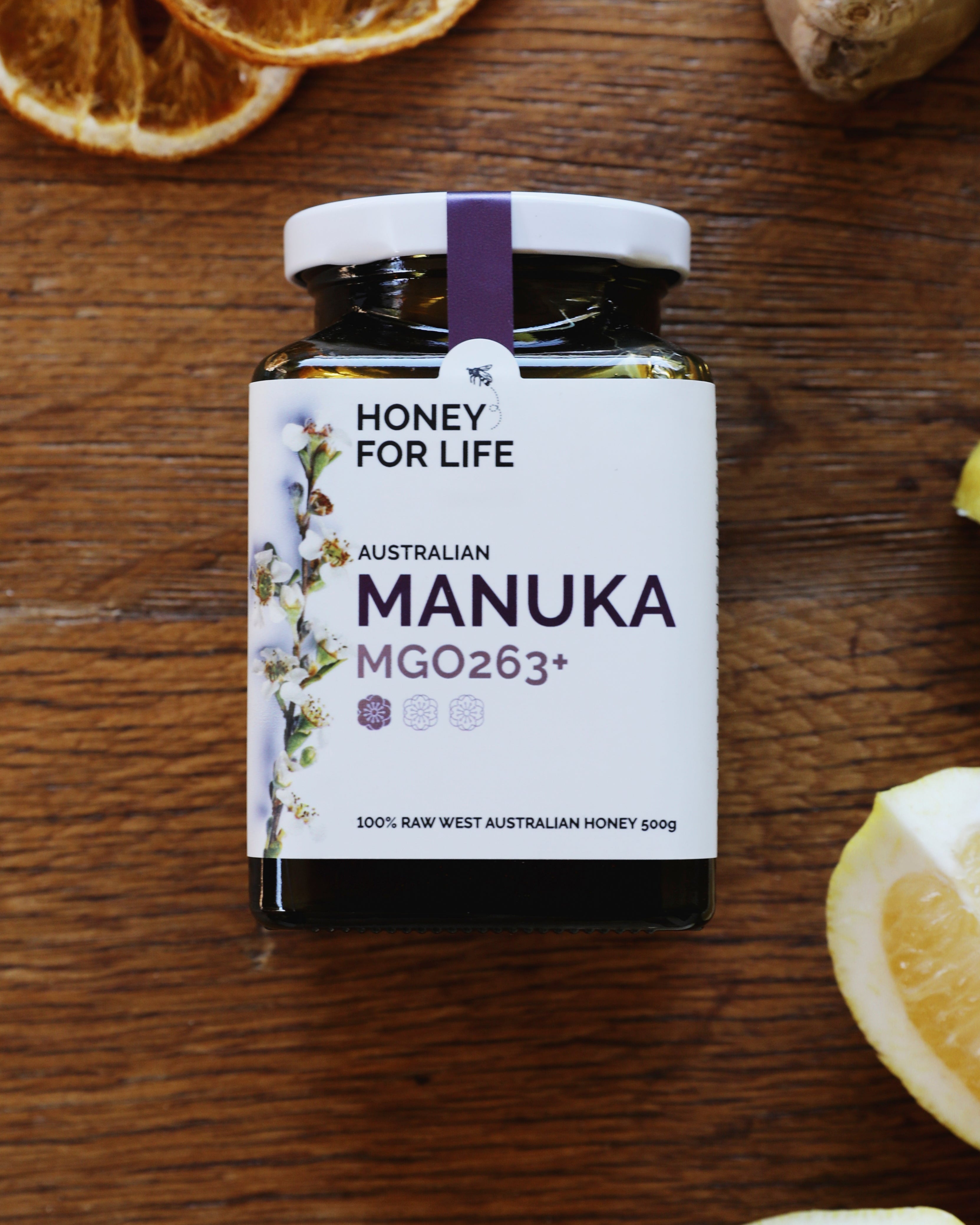 A jar of Australian Manuka honey labeled 'HONEY FOR LIFE' with MGO263+ indicated on the label. The jar is accompanied by slices of lemon and ginger on a wooden surface.