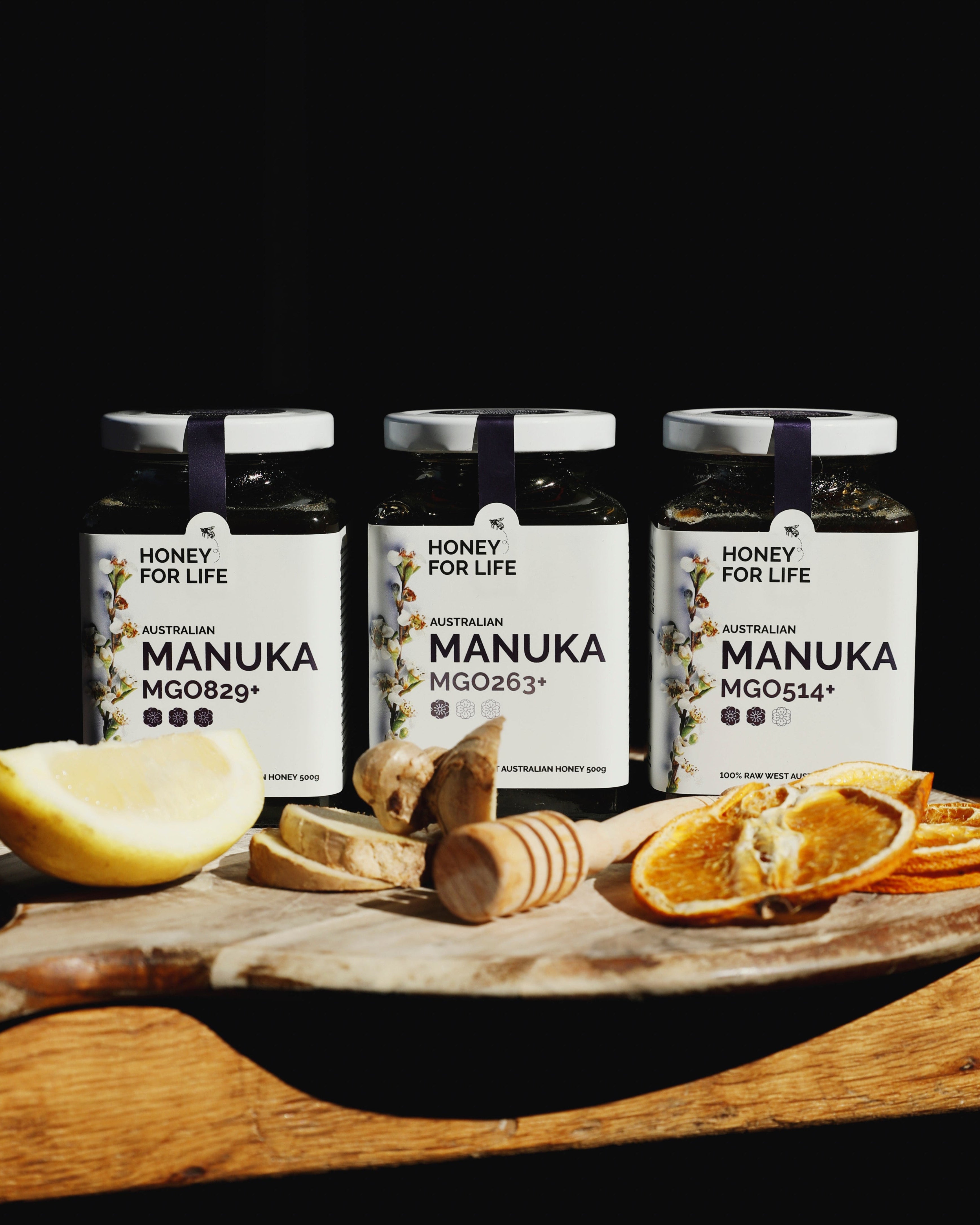 Three jars of MGO-certified Manuka honey on a wooden surface, with each jar labeled with different Manuka honey grades (MGO263+, MGO514+, MGO829+). There are also slices of lemon and manuka plant leaves in the image.