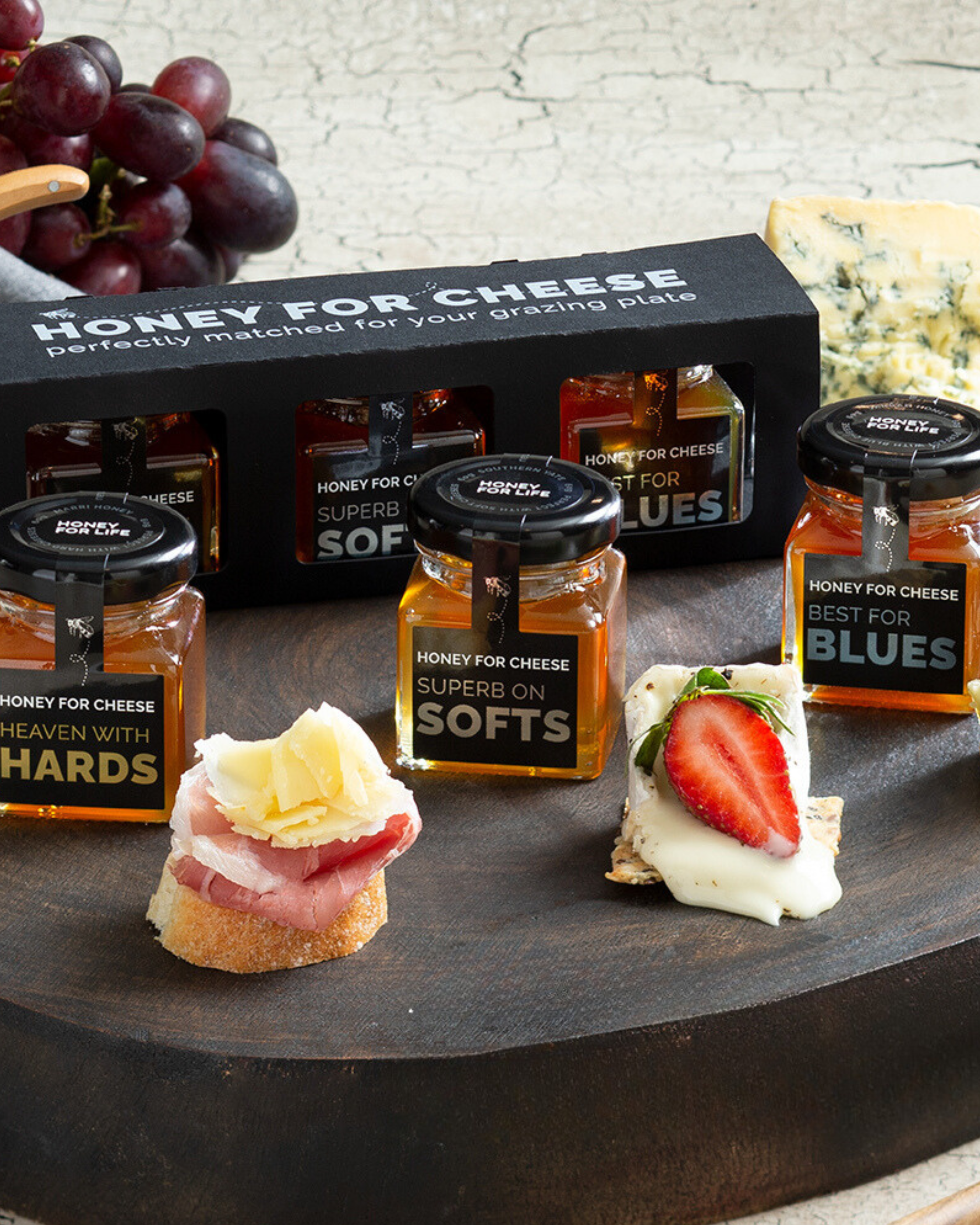 An assortment of cheeses paired with jars of honey, displayed on a wooden board with a black packaging box in the background. Cheeses are served with slices of fruit and meats, indicating a charcuterie board setup.