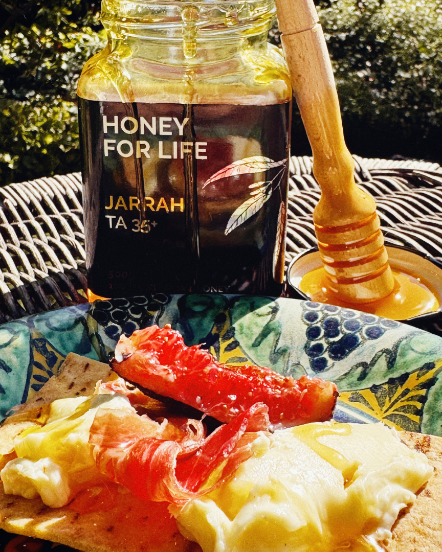 Honey jar labeled 'Honey for Life' with a honey dipper on a plate with food outdoors.